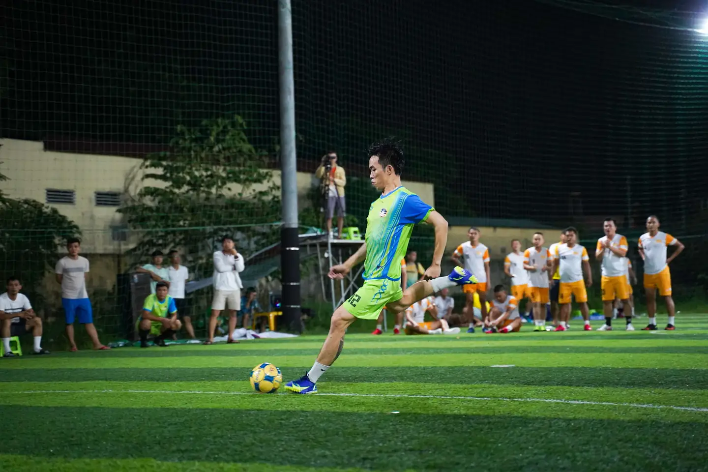Partido de fútbol de noche en una cancha sintética con malla: un jugador cobra mientras los demás esperan.